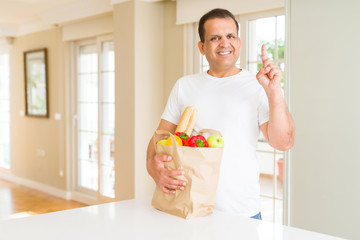 Middle age man holding groceries shopping bag at home surprised with an idea or question pointing finger with happy face, number one