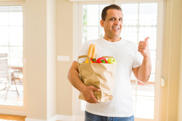 Middle age man holding groceries shopping bag at home pointing and showing with thumb up to the side with happy face smiling