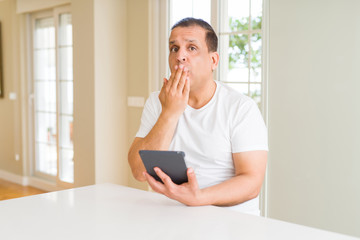 Middle age man using digital table at home cover mouth with hand shocked with shame for mistake, expression of fear, scared in silence, secret concept