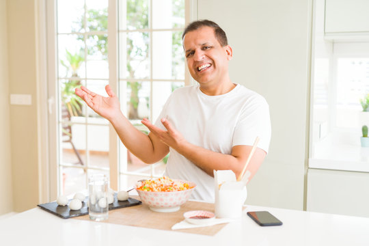 Middle age man eating asian food with chopsticks at home amazed and smiling to the camera while presenting with hand and pointing with finger.