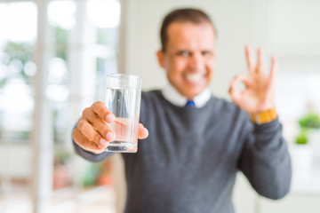 Middle age man drinking glass of water at home doing ok sign with fingers, excellent symbol