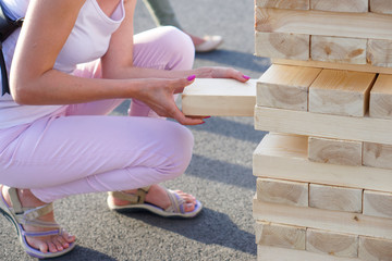 A girl in pink takes out one bar from the tower while playing a giant jenga. Outside