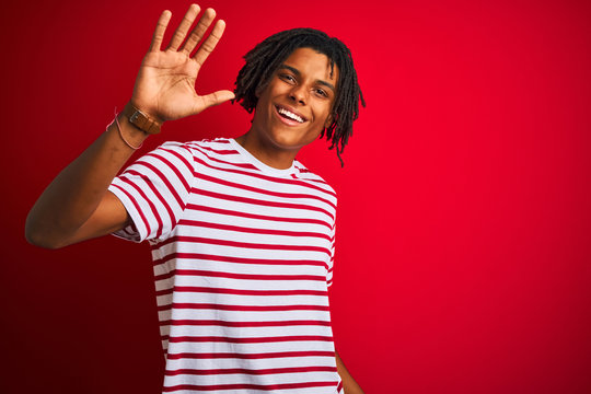 Young afro man with dreadlocks wearing striped t-shirt standing over isolated red background Waiving saying hello happy and smiling, friendly welcome gesture