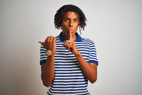 Afro man with dreadlocks wearing striped blue polo standing over isolated white background asking to be quiet with finger on lips pointing with hand to the side. Silence and secret concept.