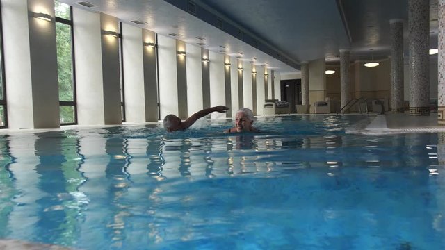 Happy mature woman and man conversating while swimming in clear blue water of indoor pool of luxury spa hotel. Joyful loving elderly couple on holidays, healthy lifestyle, wealthy retirement concept