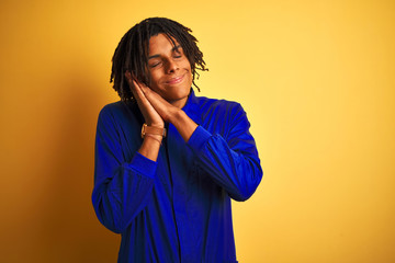 Afro worker man with dreadlocks wearing mechanic uniform over isolated yellow background sleeping tired dreaming and posing with hands together while smiling with closed eyes.