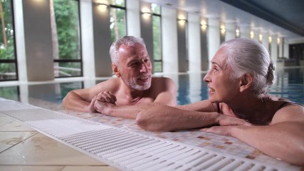 Close-up of cheerful elderly gray-haired husband and wife laughing enjoying conversation in swimming pool of luxury spa hotel. Loving aging couple having fun on vacation, positive lifestyle in old age