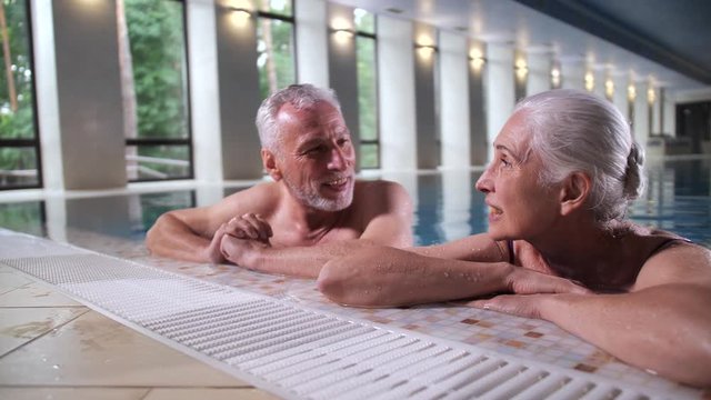 Close-up of cheerful elderly gray-haired husband and wife laughing enjoying conversation in swimming pool of luxury spa hotel. Loving aging couple having fun on vacation, positive lifestyle in old age