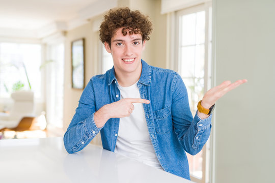 Young handsome man wearing casual denim jacket at home amazed and smiling to the camera while presenting with hand and pointing with finger.