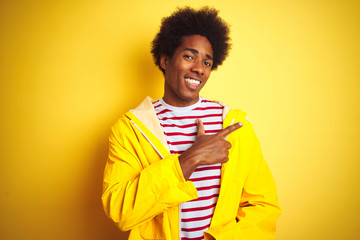 African american man with afro hair wearing rain coat standing over isolated yellow background cheerful with a smile of face pointing with hand and finger up to the side 