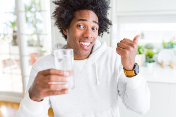 African American man drinking a glass of water at home pointing and showing with thumb up to the side with happy face smiling