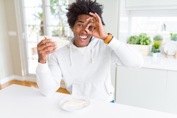 Fototapeta na wymiar African American man eating handmade sandwich at home with happy face smiling doing ok sign with hand on eye looking through fingers