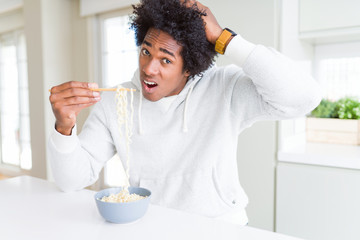 Fototapeta na wymiar African American man eating asian noodles using chopsticks at home stressed with hand on head, shocked with shame and surprise face, angry and frustrated. Fear and upset for mistake.