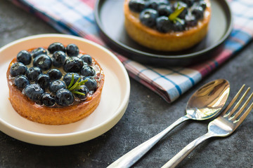 Delicious blueberry tartalets on black and white plates on a cage napkin with a spoon and fork on a grey background. Top view.
