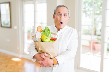 Handsome senior man holding a paper bag of fresh groceries from the supermarket scared in shock with a surprise face, afraid and excited with fear expression