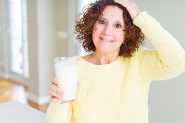 Senior woman drinking a glass of fresh milk stressed with hand on head, shocked with shame and...