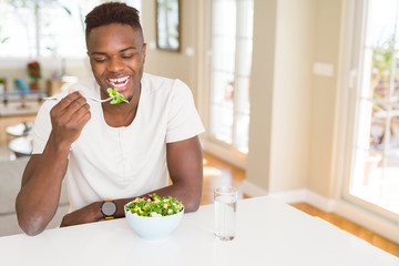 Handsome african young man eating a healthy vegetable salad using a fork to eat lettuce, happy and...