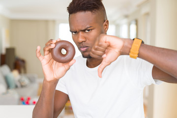African american man eating chocolate donut with angry face, negative sign showing dislike with...