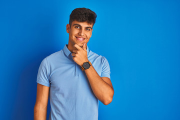 Young indian man wearing casual polo standing over isolated blue background looking confident at the camera smiling with crossed arms and hand raised on chin. Thinking positive.