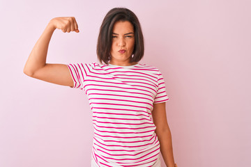 Young beautiful woman wearing striped t-shirt standing over isolated pink background Strong person showing arm muscle, confident and proud of power