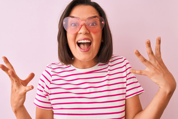 Young beautiful woman wearing fashion sunglasses with hearts over isolated pink background very happy and excited, winner expression celebrating victory screaming with big smile and raised hands