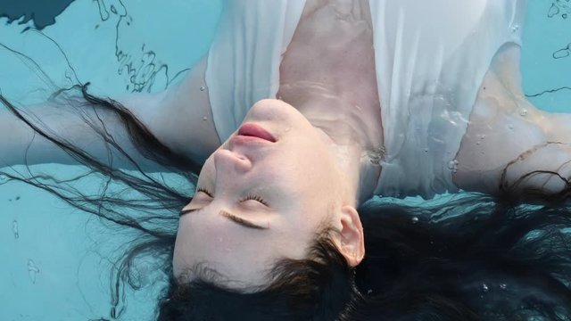 Portrait of a young brunette woman. The girl lies in the pool with her eyes closed. Relaxing spa treatments. Model in a white bikini.