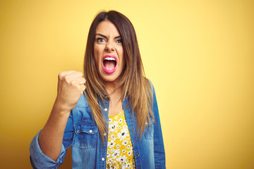 Young beautiful woman standing over yellow isolated background angry and mad raising fist frustrated and furious while shouting with anger. Rage and aggressive concept.