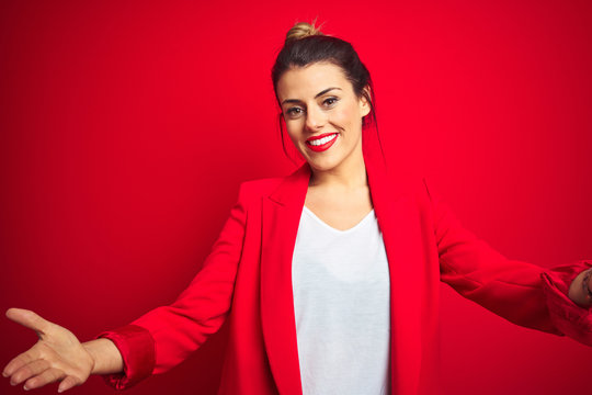 Young beautiful business woman standing over red isolated background looking at the camera smiling with open arms for hug. Cheerful expression embracing happiness.
