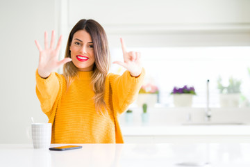 Young beautiful woman drinking a cup of coffee at home showing and pointing up with fingers number...