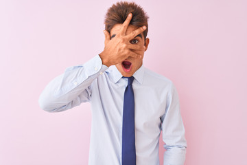 Young handsome businessman wearing shirt and tie standing over isolated pink background peeking in shock covering face and eyes with hand, looking through fingers with embarrassed expression.