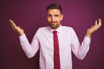 Young business man wearing elegant shirt and tie over purple isolated background clueless and confused expression with arms and hands raised. Doubt concept.