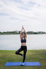 Fototapeta premium Slim beautiful women practices yoga poses on the mat beside a lake at the outdoor park with green grass background. Yoga and life health concept with copy and paste space.
