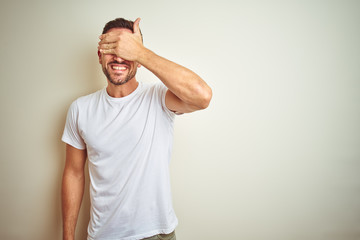 Young handsome man wearing casual white t-shirt over isolated background smiling and laughing with hand on face covering eyes for surprise. Blind concept.