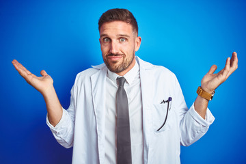 Young handsome doctor man wearing white professional coat over isolated background clueless and confused expression with arms and hands raised. Doubt concept.