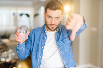 Young handsome man drinking a glass of water at home with angry face, negative sign showing dislike...