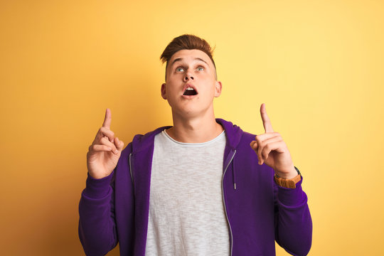 Young handsome man wearing purple sweatshirt standing over isolated yellow background amazed and surprised looking up and pointing with fingers and raised arms.