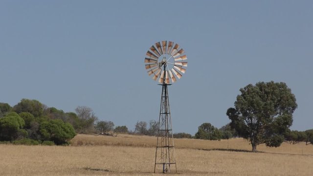 Iconic spinning wind mill on open field in outback Australia, with sunny blue sky and horizon as background.
