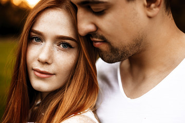 Close up portrait of a beautiful caucasian young couple embracing while she is looking at camera smiling and he is smelling her hair with closed eyes outside.