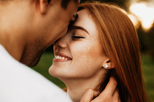 Close up side view portrait of a lovely female with freckles and red hair smiling with closed eyes before kissing while her boyfriend is touching her hair