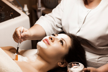 Side view portrait of a beautiful brunette leaning on a spa bed with eyes opened while doing a white skin care mask on her face and neck in a wellness spa center.