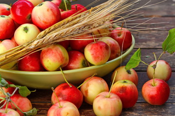 Deep bowl with red yellow apples with green foliage, scattered apples around it and bunch of ears of ripe wheat grains on old wooden plank surface. Close up