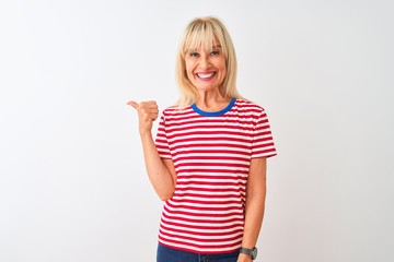 Middle age woman wearing casual striped t-shirt standing over isolated white background smiling with happy face looking and pointing to the side with thumb up.