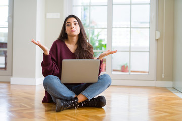 Young woman using computer laptop sitting on the floor clueless and confused expression with arms...