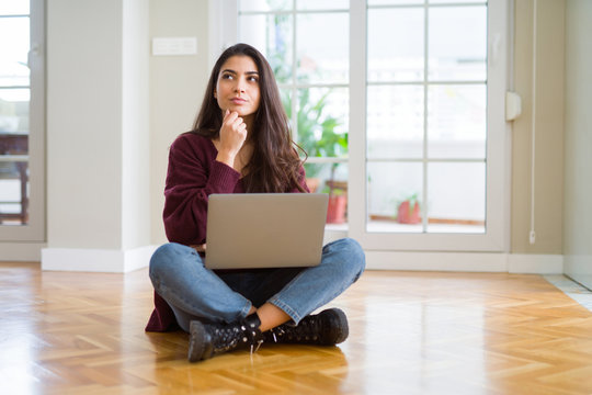 Young woman using computer laptop sitting on the floor with hand on chin thinking about question, pensive expression. Smiling with thoughtful face. Doubt concept.