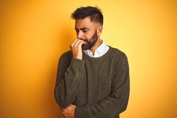 Young indian man wearing green sweater and shirt standing over isolated yellow background looking stressed and nervous with hands on mouth biting nails. Anxiety problem.