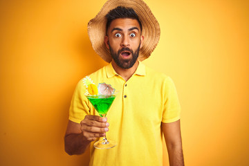 Indian man on vacation drinking cocktail wearing summer hat over isolated yellow background scared...