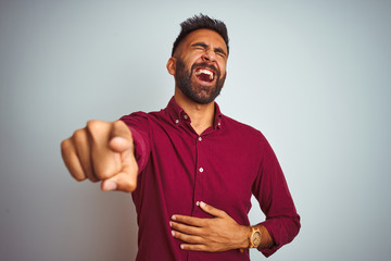 Young indian man wearing red elegant shirt standing over isolated grey background laughing at you, pointing finger to the camera with hand over body, shame expression