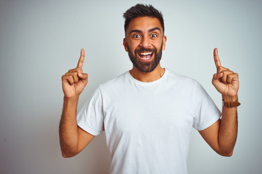 Young indian man wearing t-shirt standing over isolated white background smiling amazed and surprised and pointing up with fingers and raised arms.