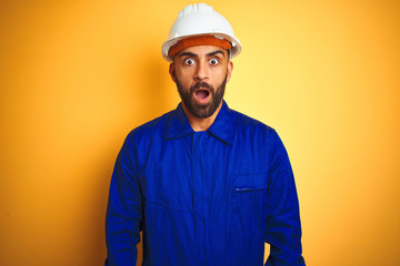 Handsome indian worker man wearing uniform and helmet over isolated yellow background afraid and...