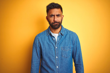 Young indian man wearing denim shirt standing over isolated yellow background skeptic and nervous, frowning upset because of problem. Negative person.